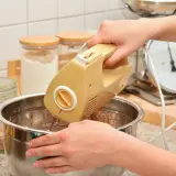 Ręce mieszają ciasto żółtym mikserem. W tle słoiki z mąką i waga kuchenna. Idealny sprzęt do kuchni, warto sprawdzić ranking mikserów ręcznych.