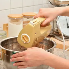 Ręce mieszają ciasto żółtym mikserem. W tle słoiki z mąką i waga kuchenna. Idealny sprzęt do kuchni, warto sprawdzić ranking mikserów ręcznych.