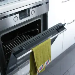Otwarty piekarnik z rusztem i naczyniem, na drzwiczkach wisi zielony ręcznik. To przygotowanie do pyrolizy w piekarniku.
