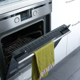 Otwarty piekarnik z rusztem i naczyniem, na drzwiczkach wisi zielony ręcznik. To przygotowanie do pyrolizy w piekarniku.