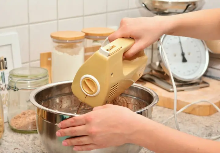 Ręce mieszają ciasto żółtym mikserem. W tle słoiki z mąką i waga kuchenna. Idealny sprzęt do kuchni, warto sprawdzić ranking mikserów ręcznych.