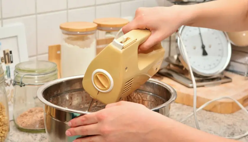 Ręce mieszają ciasto żółtym mikserem. W tle słoiki z mąką i waga kuchenna. Idealny sprzęt do kuchni, warto sprawdzić ranking mikserów ręcznych.