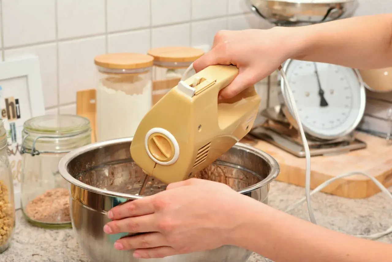 Ręce mieszają ciasto żółtym mikserem. W tle słoiki z mąką i waga kuchenna. Idealny sprzęt do kuchni, warto sprawdzić ranking mikserów ręcznych.